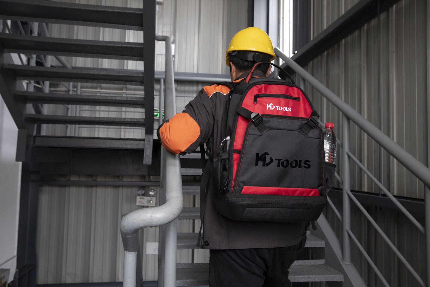 Mit einem gelben Schutzhelm und einer orange-schwarzen Jacke trägt eine Person einen HC Tools Premium Werkzeugrucksack in Schwarz und Rot, während sie eine Metalltreppe in einem Industriegebäude hinaufgeht.