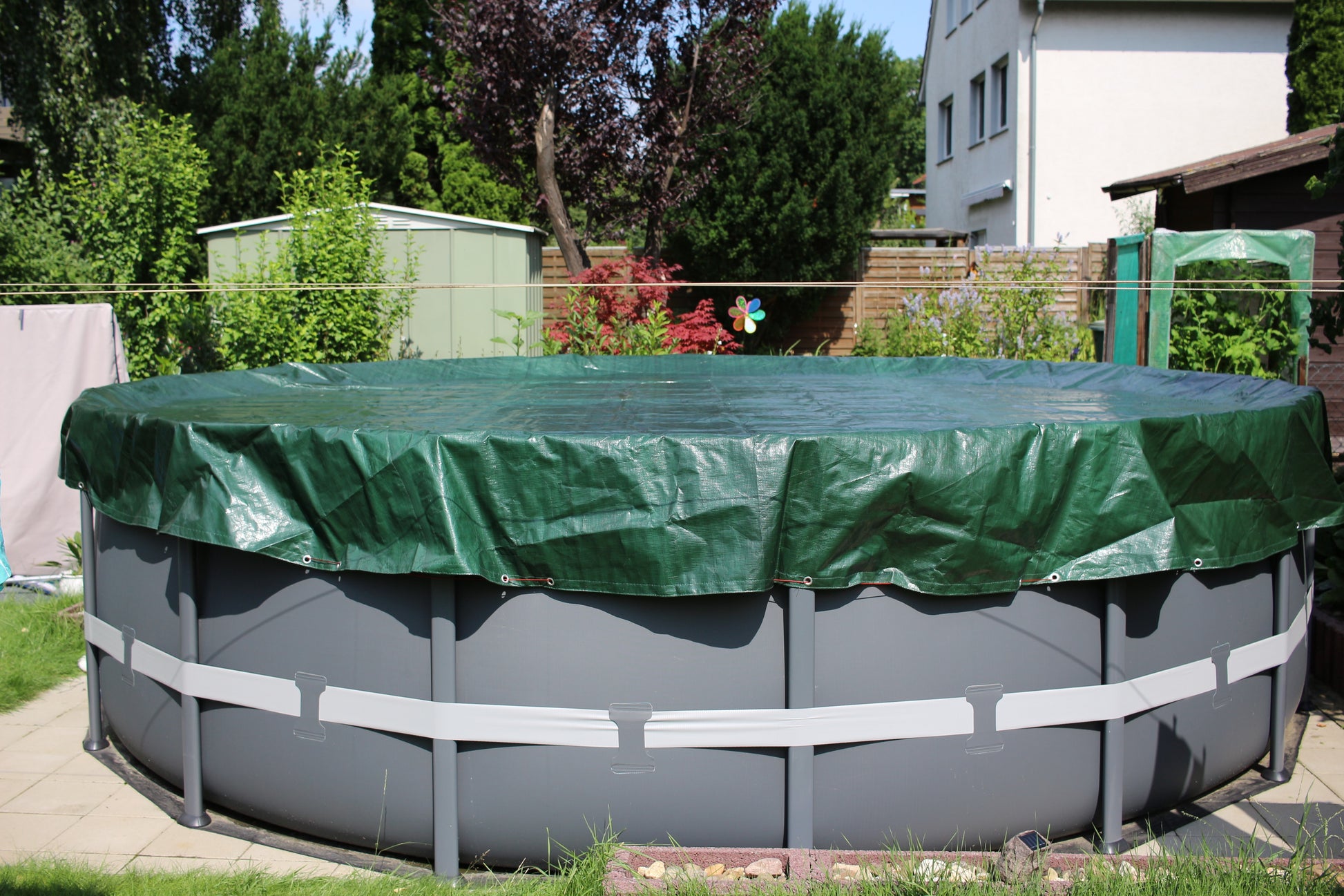 Ein großer runder oberirdischer Pool ist mit der HC Garten & Freizeit Sommer- & Winter-Abdeckplane für runde Pools 8,0 m abgedeckt. Der Pool liegt in einem grasbewachsenen Garten mit Bäumen, Gartenhäusern und einem weißen Haus im Hintergrund.