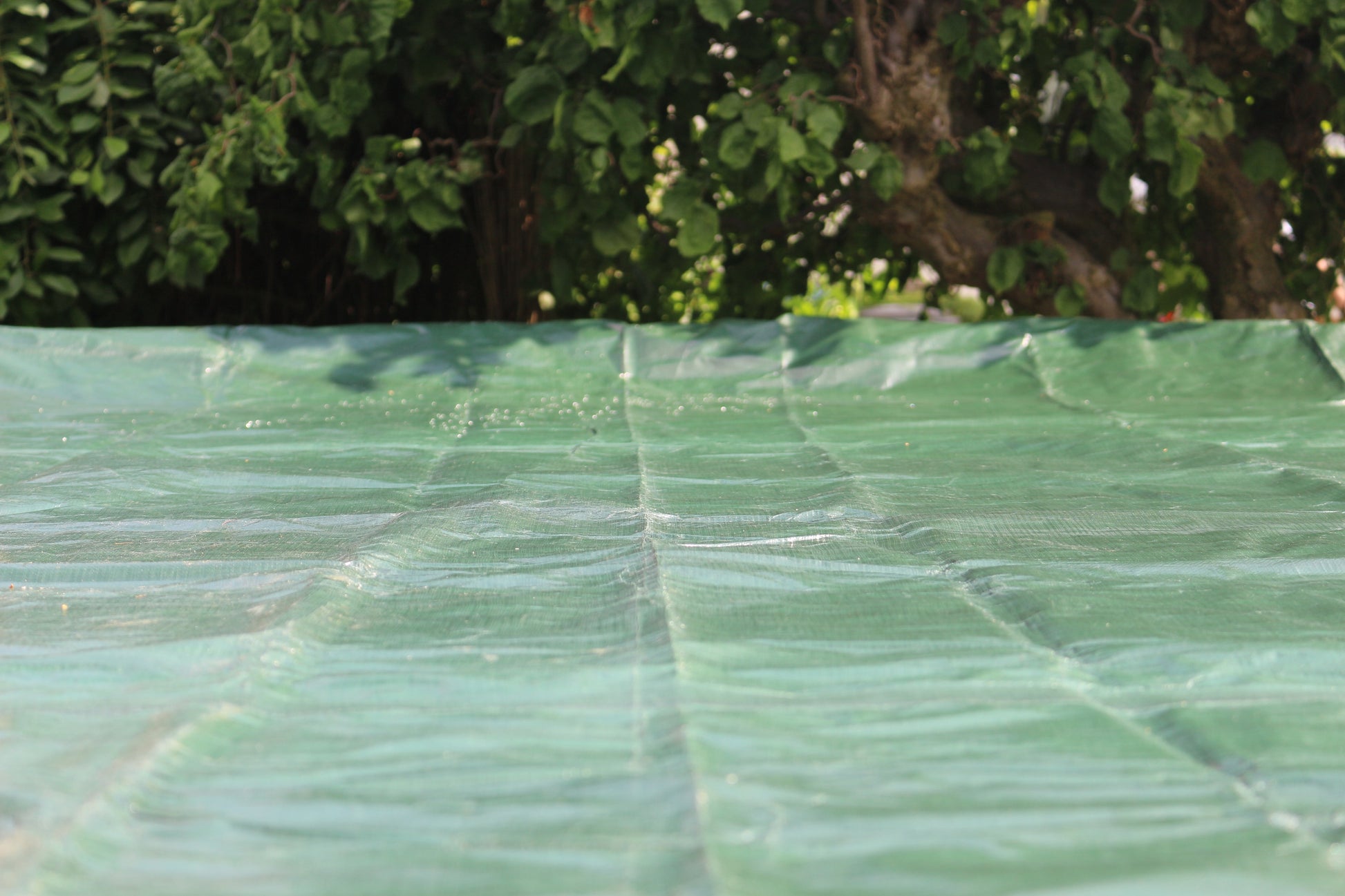 Eine Nahaufnahme der HC Garten & Freizeit Sommer- & Winter-Abdeckplane für runde Pools 5,0 m, flach ausgebreitet mit belaubten Bäumen dahinter. Die Oberfläche der Polyethylen-Folie wird im Detail gezeigt, wobei ihre Struktur und Falten hervorgehoben werden.