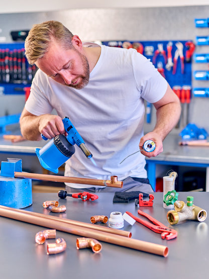 Ein Mann in einem weißen T-Shirt bearbeitet mit der CFH Lötlampe PZO 7000 - 52192 von CFH Löt- und Gasgeräte GmbH und Lötzinn ein Kupferrohr an einer Werkbank, umgeben von Klempnerwerkzeugen und Kupferfittings in einer Werkstatt.