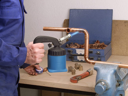 Eine Person in einem blauen Arbeitsanzug benutzt die CFH Lötlampe LM 2000 der CFH Löt- und Gasgeräte GmbH zum Löten von Kupferrohren auf einer Werkbank, wobei im Hintergrund Werkzeuge, Fittings und ein blauer Werkzeugkasten zu sehen sind.