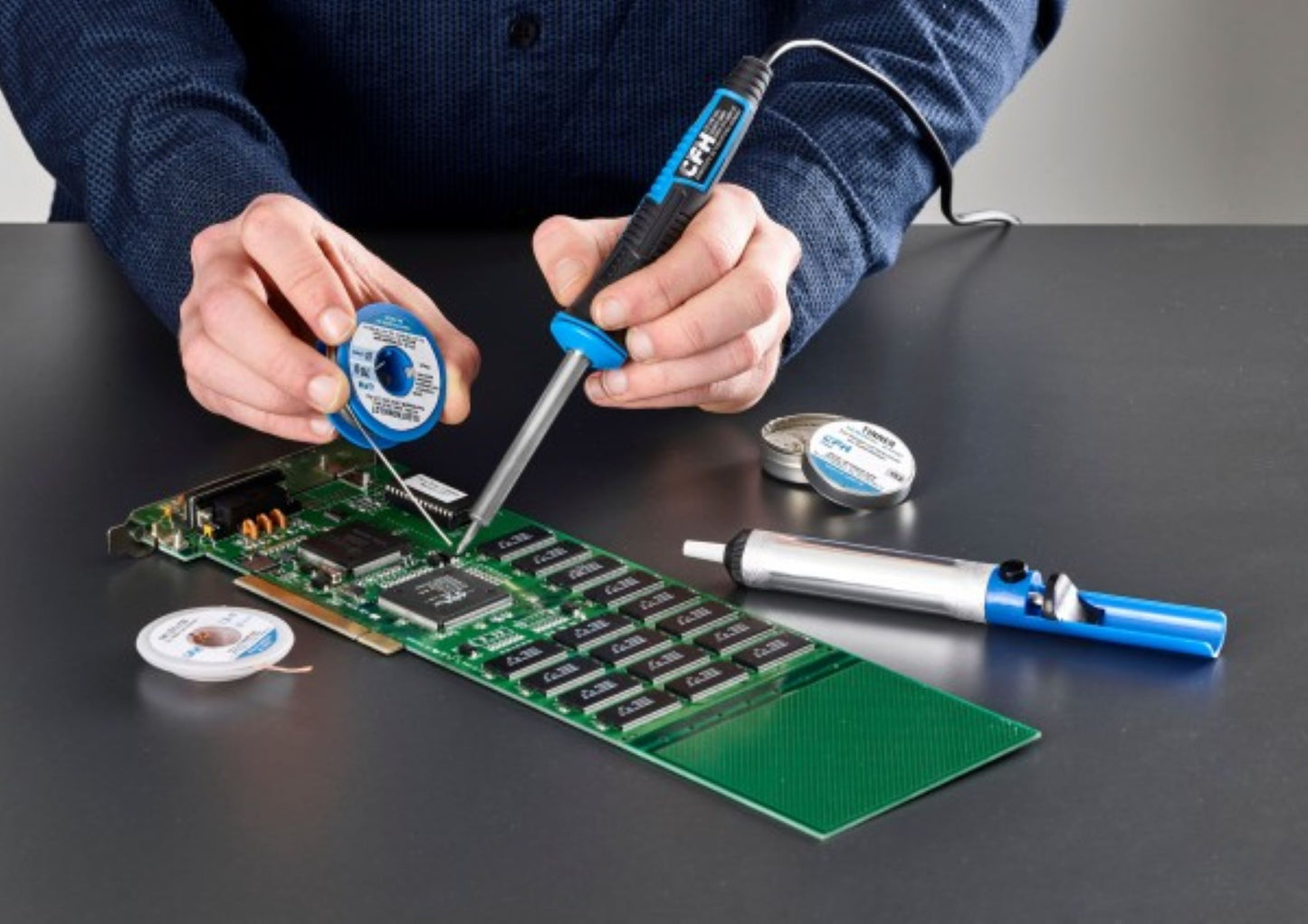 Eine Person lötet mit einem CFH Elektrolötkolben E 15 der CFH Löt- und Gasgeräte GmbH und Lötdraht Bauteile auf eine grüne Leiterplatte. Auf dem grauen Tisch liegen zusätzliche Lötspulen, eine Entlötpumpe und eine Lötspitze.