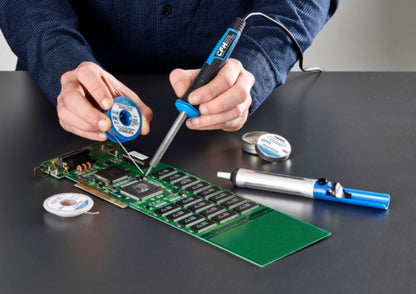 Eine Person lötet mit einem CFH Elektrolötkolben E 15 der CFH Löt- und Gasgeräte GmbH und Lötdraht Bauteile auf eine grüne Leiterplatte. Auf dem grauen Tisch liegen zusätzliche Lötspulen, eine Entlötpumpe und eine Lötspitze.