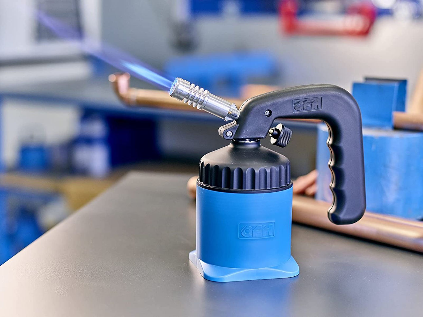 Eine blaue CFH Lötlampe LM 2000 - 521163 der CFH Löt- und Gasgeräte GmbH mit schwarzem Griff entzündet eine blaue Flamme auf einer Werkbank, Kupferrohre und Werkzeuge verschwimmen im Hintergrund der Werkstatt.