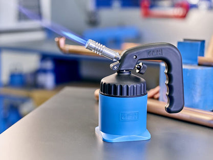 Eine blaue CFH Lötlampe LM 2000 - 521163 der CFH Löt- und Gasgeräte GmbH mit schwarzem Griff entzündet eine blaue Flamme auf einer Werkbank, Kupferrohre und Werkzeuge verschwimmen im Hintergrund der Werkstatt.