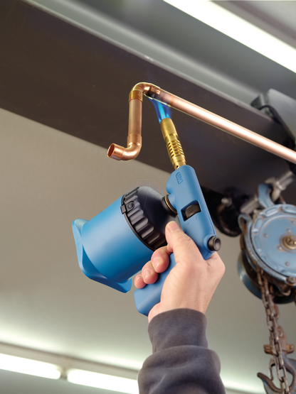 Eine Person benutzt die CFH Lötlampe PZO 7000 - 52192 der CFH Löt- und Gasgeräte GmbH, um eine Kupferrohrverbindung zum Löten zu erwärmen. Im Hintergrund sind Industrieanlagen und Beleuchtung zu sehen.