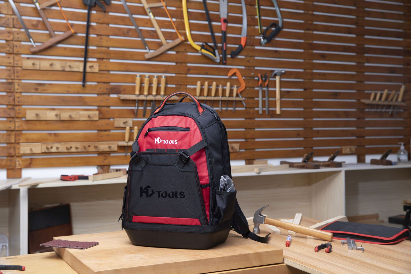 Ein schwarz-roter HC Tools Premium Werkzeugrucksack liegt auf einer hölzernen Werkbank in einer Werkstatt, umgeben von Handwerkzeugen wie einem Hammer, einem Maßband und Sägen, die an einem Wandregal hängen.