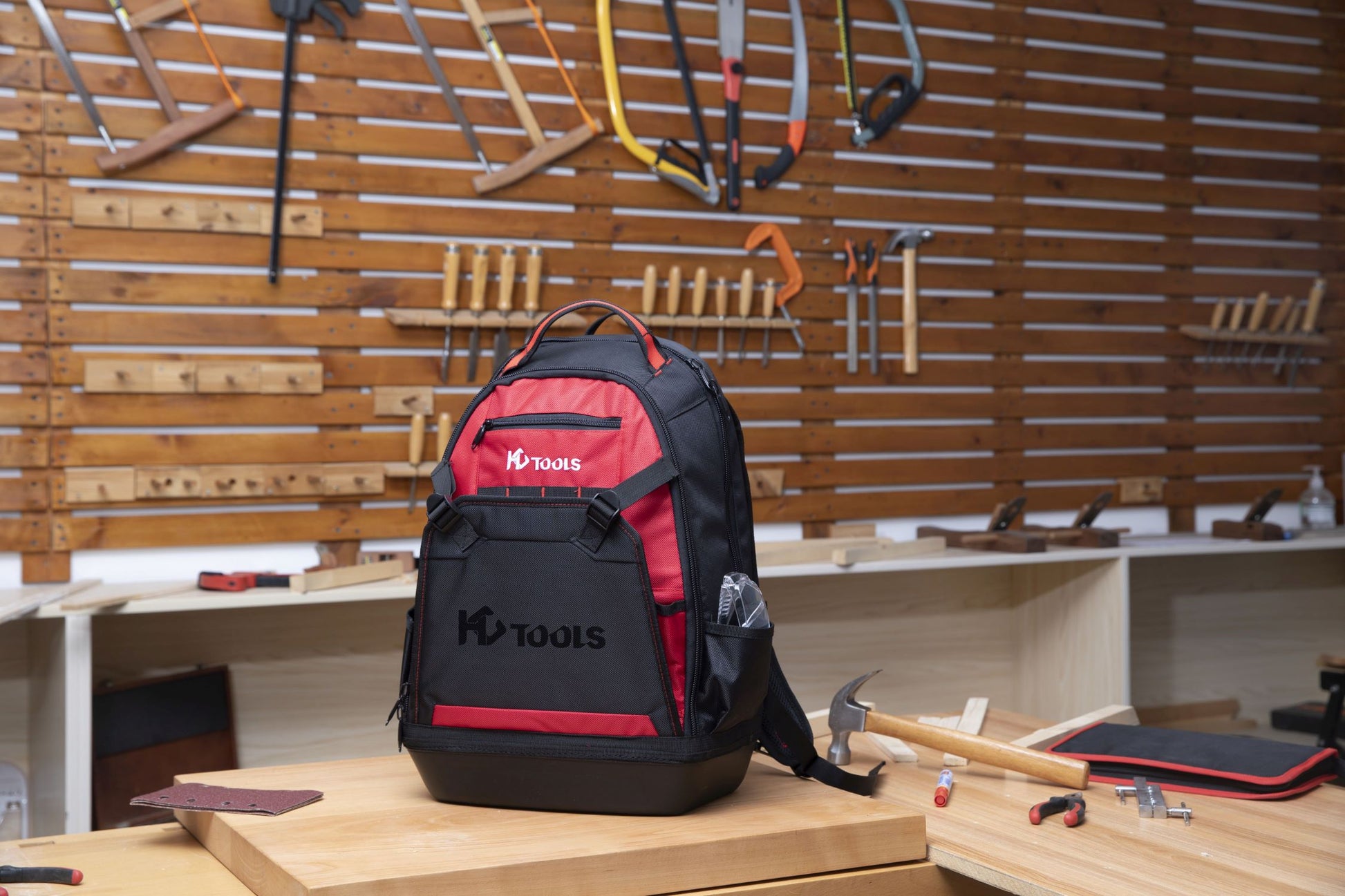 Ein schwarz-roter HC Tools Premium Werkzeugrucksack liegt auf einer hölzernen Werkbank in einer Werkstatt, umgeben von Handwerkzeugen wie einem Hammer, einem Maßband und Sägen, die an einem Wandregal hängen.