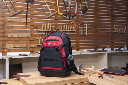 Ein schwarz-roter HC Tools Premium Werkzeugrucksack liegt auf einer hölzernen Werkbank in einer Werkstatt, umgeben von Handwerkzeugen wie einem Hammer, einem Maßband und Sägen, die an einem Wandregal hängen.