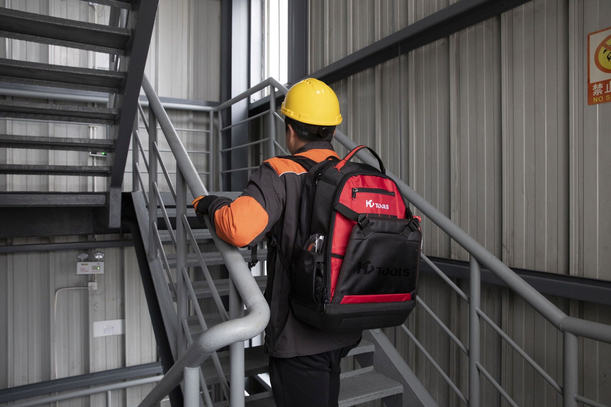 Eine Person mit einem gelben Schutzhelm und einer orange-schwarzen Uniform trägt den HC Tools Premium Werkzeugrucksack, während sie eine Metalltreppe in einem Industriegebäude hinaufgeht.